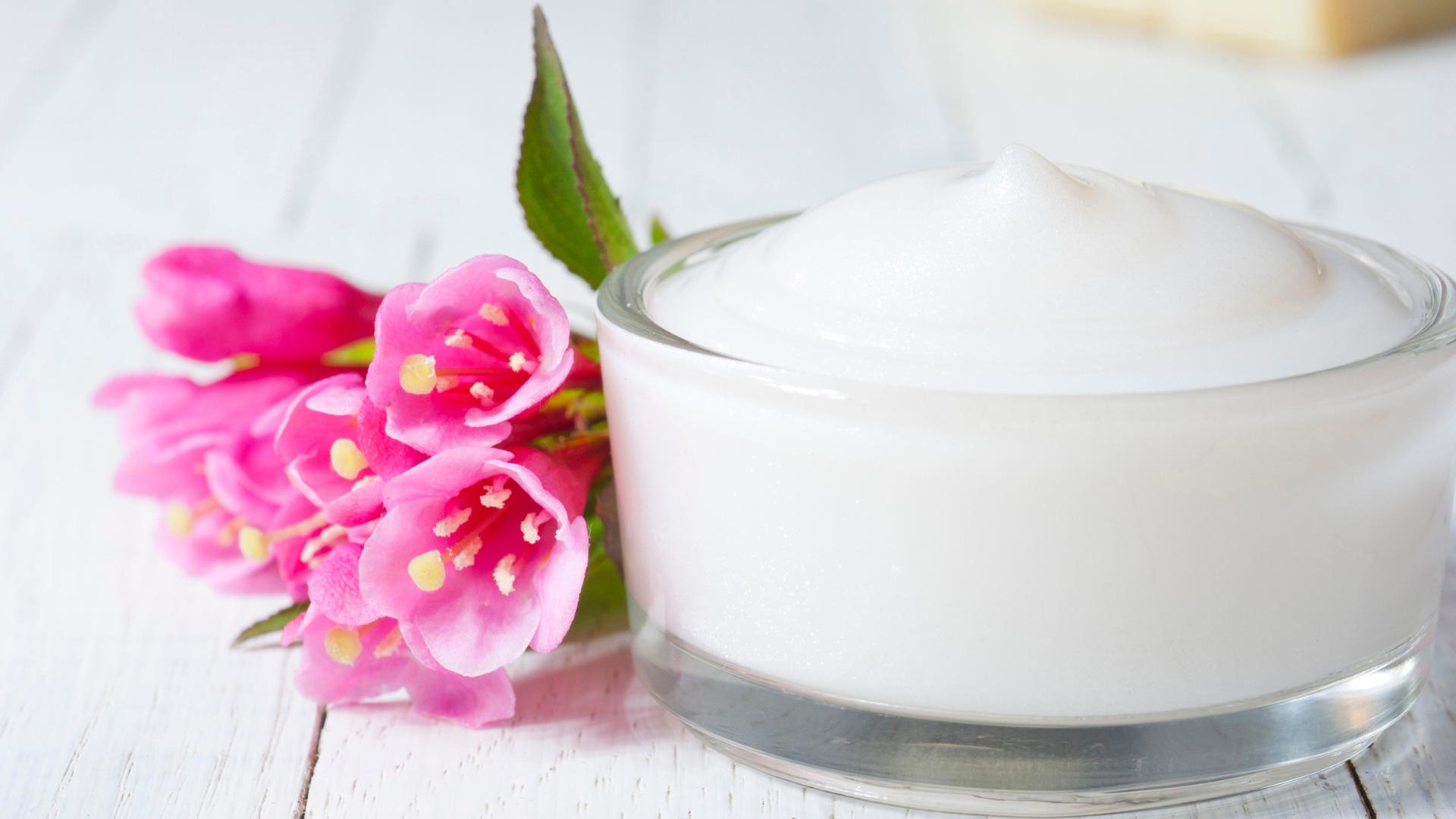 Offener Cremetiegel mit weißer Creme und rosa Blüten auf hellem Holz